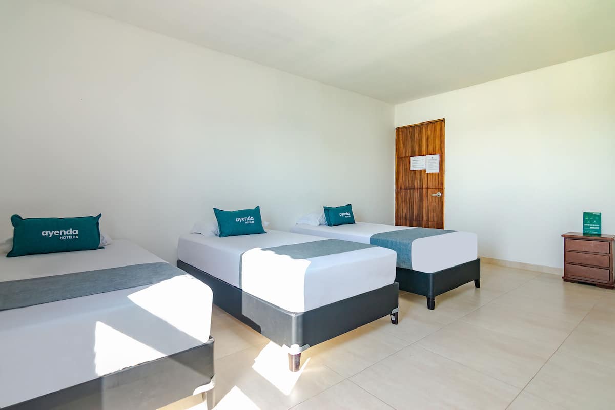 Habitación de hotel con dos camas, cada una con cubrecama gris y blanco, y almohadas color turquesa con el nombre del hotel. Se observa un armario de madera y una pequeña mesita de noche con un letrero verde.
