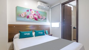 Quarto de hotel com cama, quadro de flores na parede, aparelho de ar condicionado e porta de madeira.