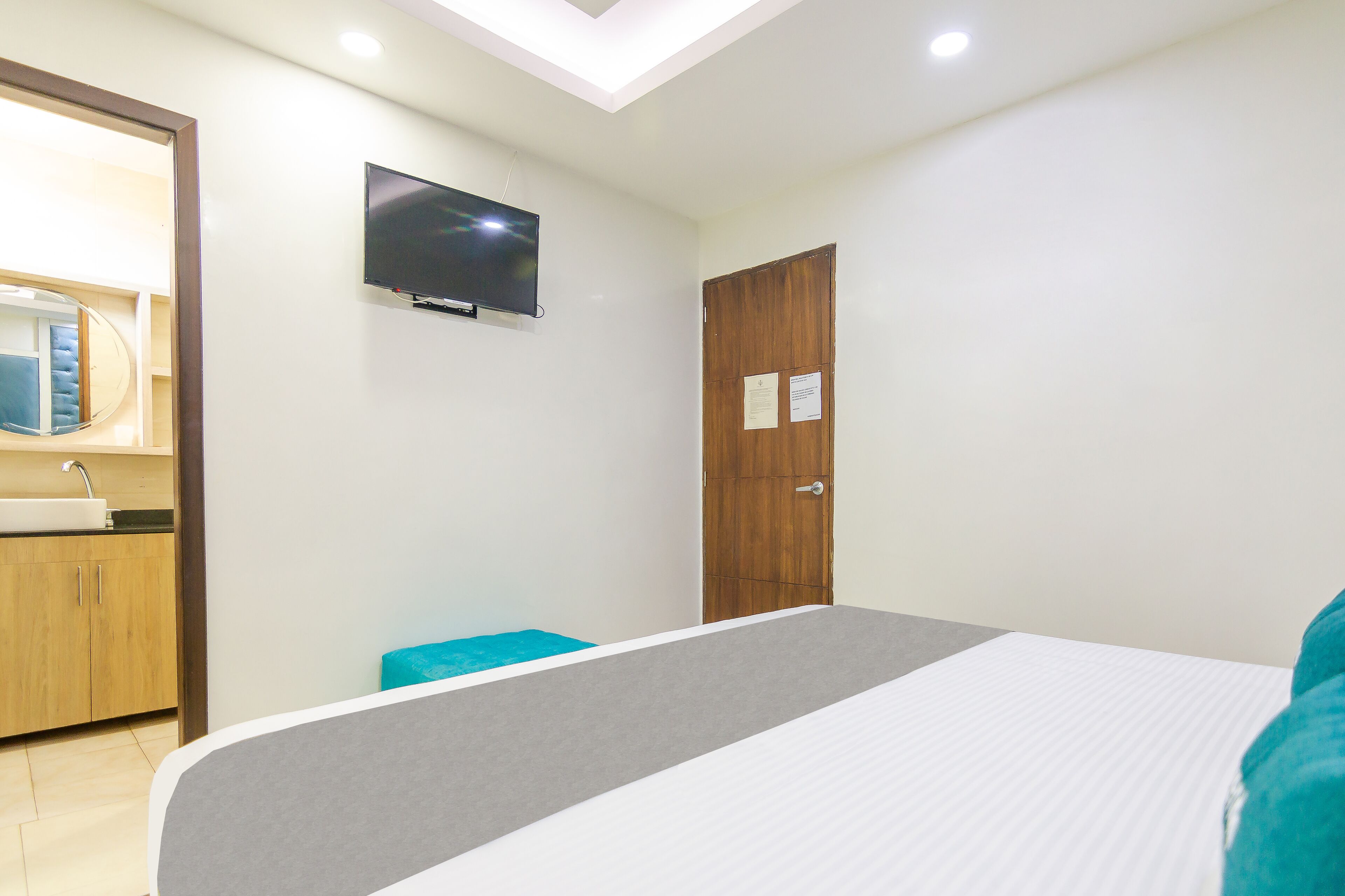 A bedroom with a bed, a television mounted on the wall, a wooden door, and a bathroom visible in the background.