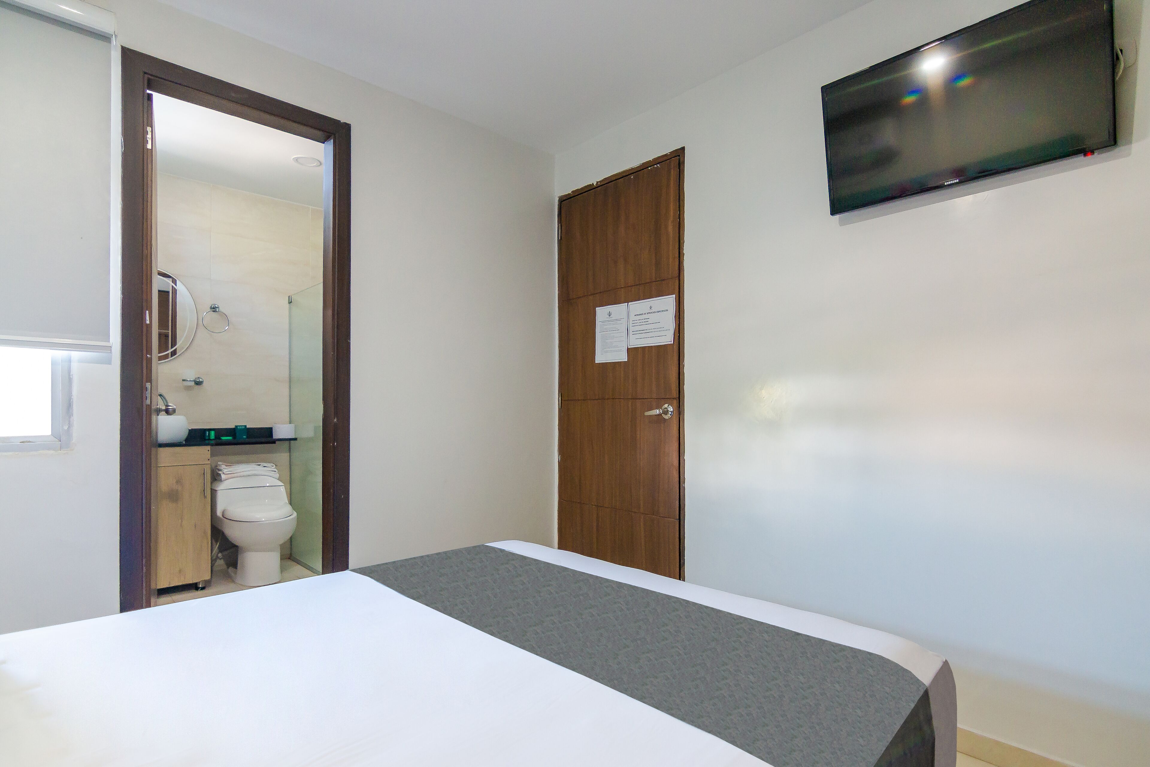 A hotel room with a bed, a wooden wardrobe, a toilet, and a TV mounted on the wall.