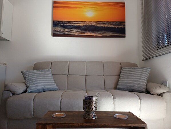 Un salon avec un canapé beige, deux coussins à rayures, une table basse en bois sur laquelle est posé un vase décoratif, et un tableau représentant un coucher de soleil accroché au mur.