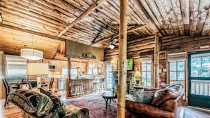 Interior rústico de una cabaña con techo de madera, comedor, sala de estar con sofá de cuero y cocina con refrigerador y microondas.