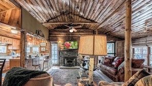 A rustic cabin interior with a stone fireplace, wooden ceiling, and a cozy seating area.