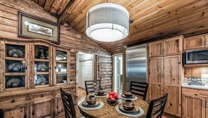 A rustic wooden cabin interior with a dining table set for four, surrounded by wooden cabinets and a large hanging light fixture. - The Apple Tree | Tranquil Rio Ruidoso Cabin w/ Deck Views, River Sounds & Cozy Charm (Ruidoso)