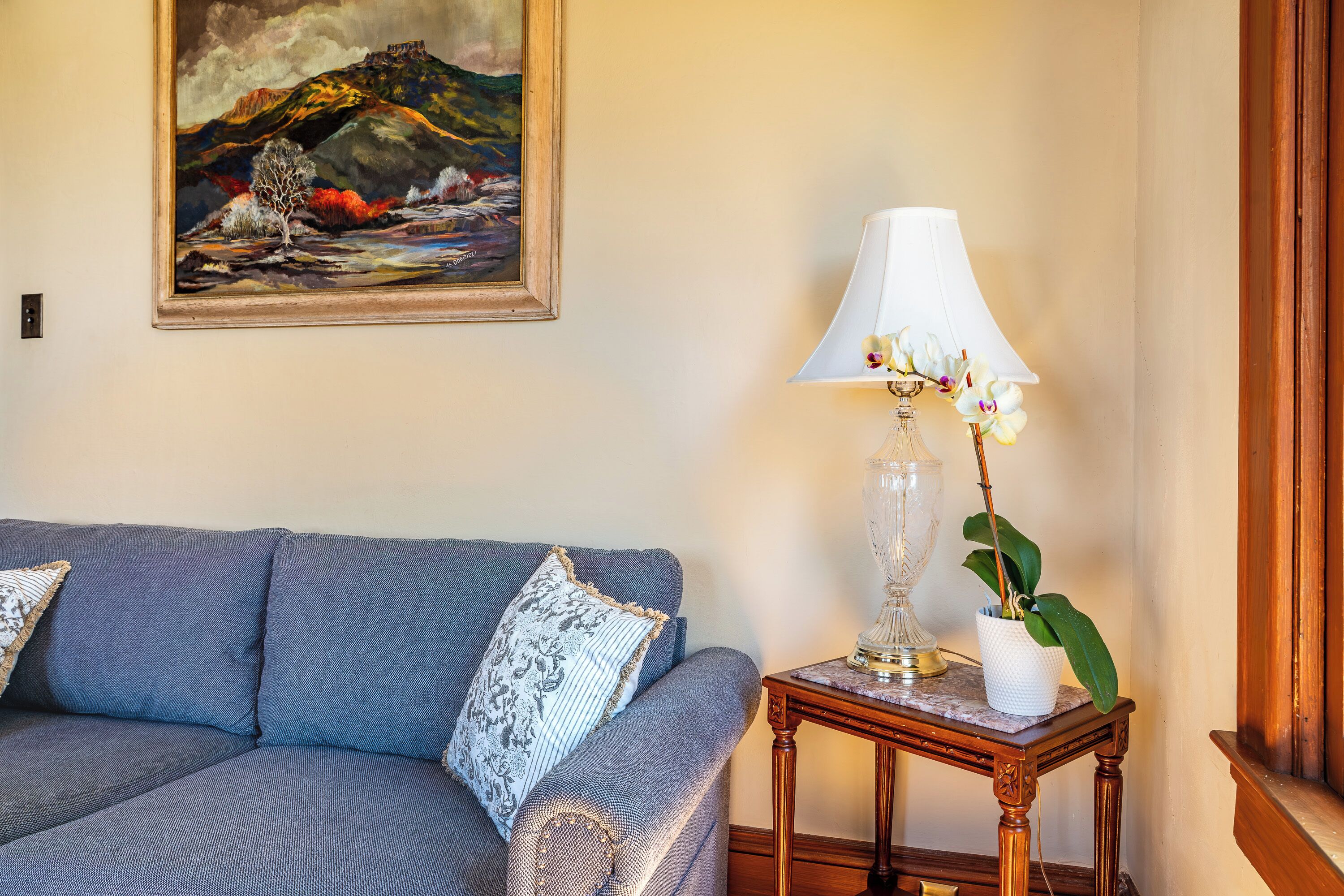 A room with a blue sofa, a framed painting, a table lamp, and a potted plant.