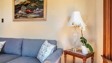 A room with a blue sofa, a framed painting, a table lamp, and a potted plant.