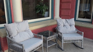 Two wicker chairs with cushions, a small table, and a potted plant on a porch with a red brick wall and windows.