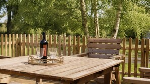 A wooden picnic table with a bottle and glasses on it, set against a wooden fence and trees. - Paddock Cottage - sleeps 2 guests in 1 bedroom (Winchcombe)