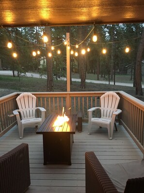 A covered deck with a central fire feature, two white Adirondack chairs, and string lights.