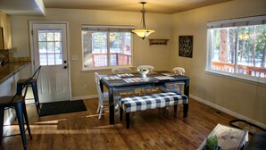 Uma área de refeições com mesa de madeira, cadeiras e um banco. Há uma porta, janelas e um lustre.