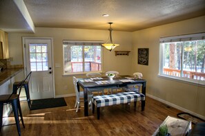 A dining area with a wooden table, chairs, and a bench. There is a door, windows, and a ceiling light fixture.
