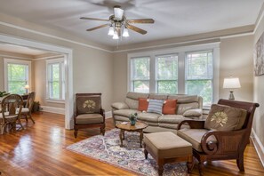 A spacious living room with a wooden floor, a beige sofa, two armchairs, a coffee table, and a ceiling fan.
