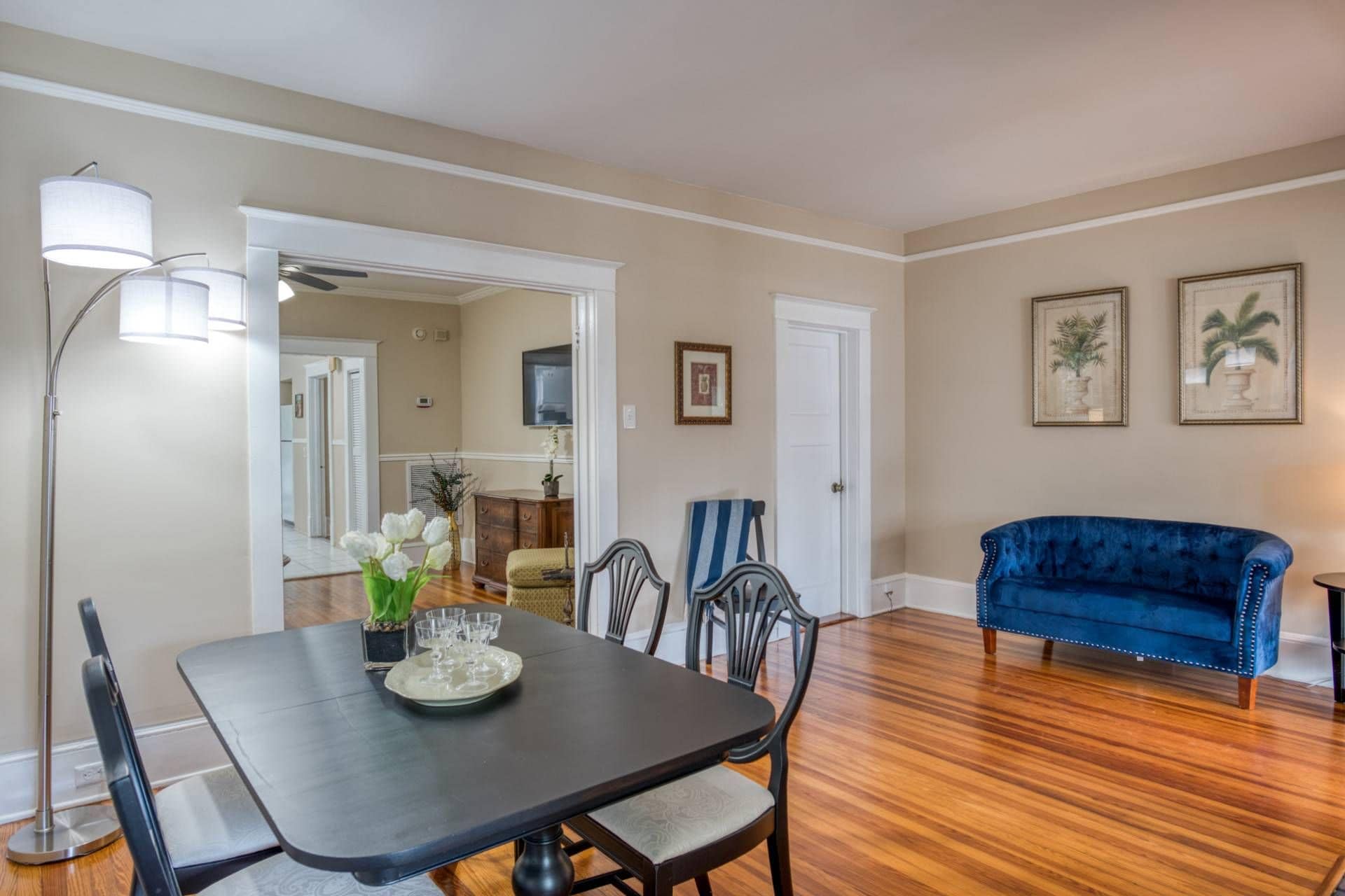 A dining area with a table, chairs, and a blue sofa. There are framed pictures on the wall and a floor lamp.