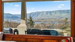 A room with a large window overlooking a scenic landscape with mountains, a river, and a small town. - Expansive Panoramic View of Columbia Gorge, Enormous Deck and Seating, BBQ, Billiards, Darts, Games (The Dalles)
