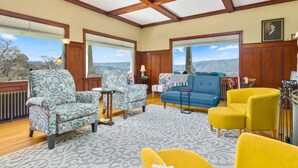 A spacious living room with a view of the countryside, featuring patterned armchairs, a blue sofa, and a yellow armchair. - Expansive Panoramic View of Columbia Gorge, Enormous Deck and Seating, BBQ, Billiards, Darts, Games (The Dalles)