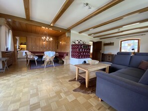 A spacious living room with wooden beams, a sofa, a dining table, and a kitchen area visible through an open door.