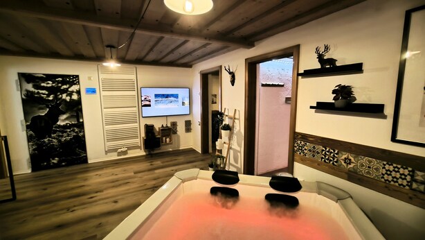 A room with a wooden floor, a large bathtub with a pink light, a mounted TV, and a deer head decoration. - Vacation home Blindauer Eck 160 sqm with mountain view (Reit im Winkl)