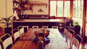Une cuisine spacieuse avec un grand îlot central, une table à manger en bois et des chaises. On y trouve des étagères remplies d’ustensiles de cuisine et de plantes.