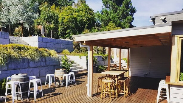 Une terrasse en bois dotée d’un espace salon couvert, d’un escalier en pierre et offrant une vue sur des arbres et un bâtiment.