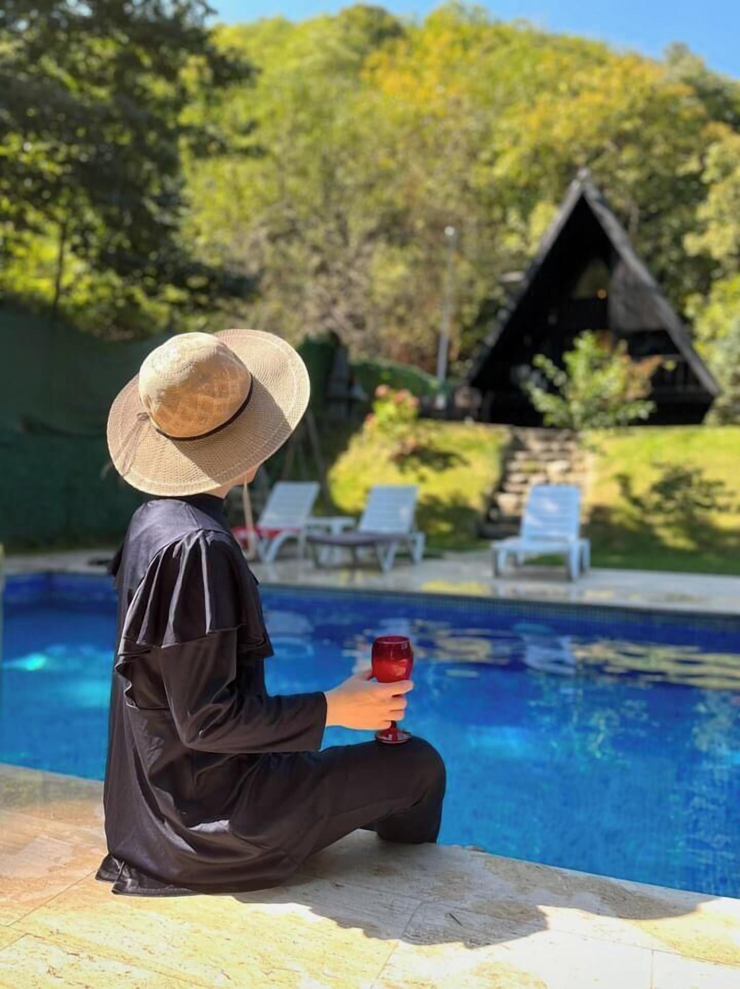 Una persona sentada junto a una piscina, sosteniendo una bebida, con sombrero y un atuendo negro.