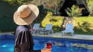 Una persona sentada junto a una piscina, sosteniendo una bebida, con sombrero y un atuendo negro.