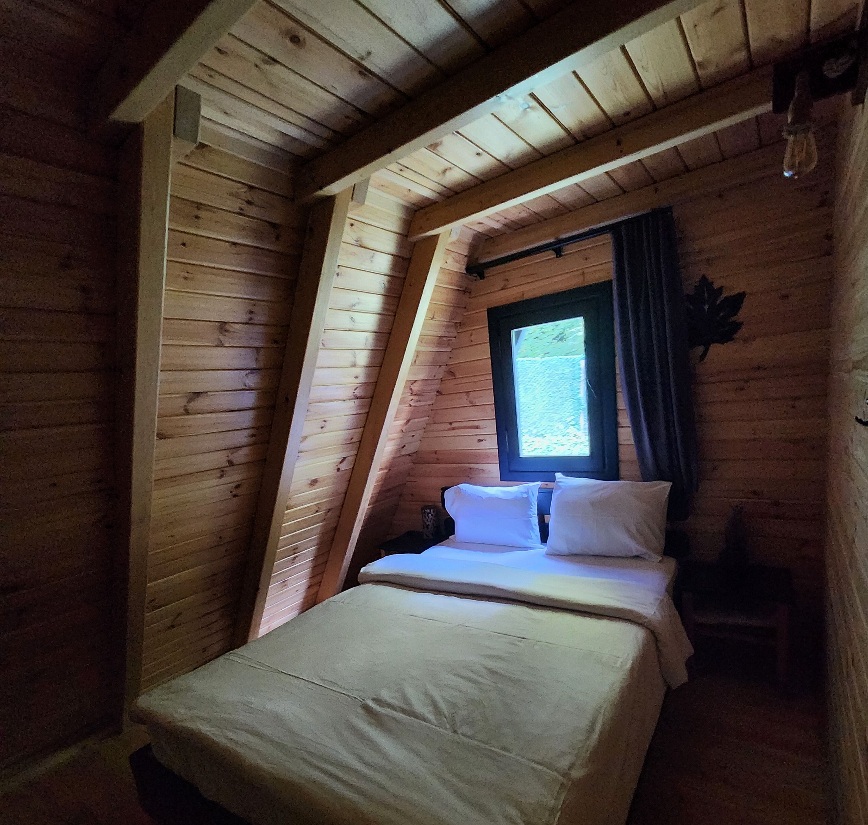 A small, cozy room with a sloped ceiling, a single bed with white bedding, a window with a view of greenery, and wooden walls.