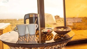 Una mesa con café, dos tazas, un tazón de cereal y una bandeja, con vista a un paisaje desértico a través de amplias ventanas.