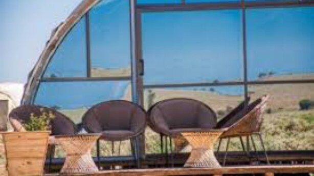Una estructura de vidrio curvada con tres sillas de mimbre y una pequeña mesa, probablemente un patio con vistas al paisaje.
