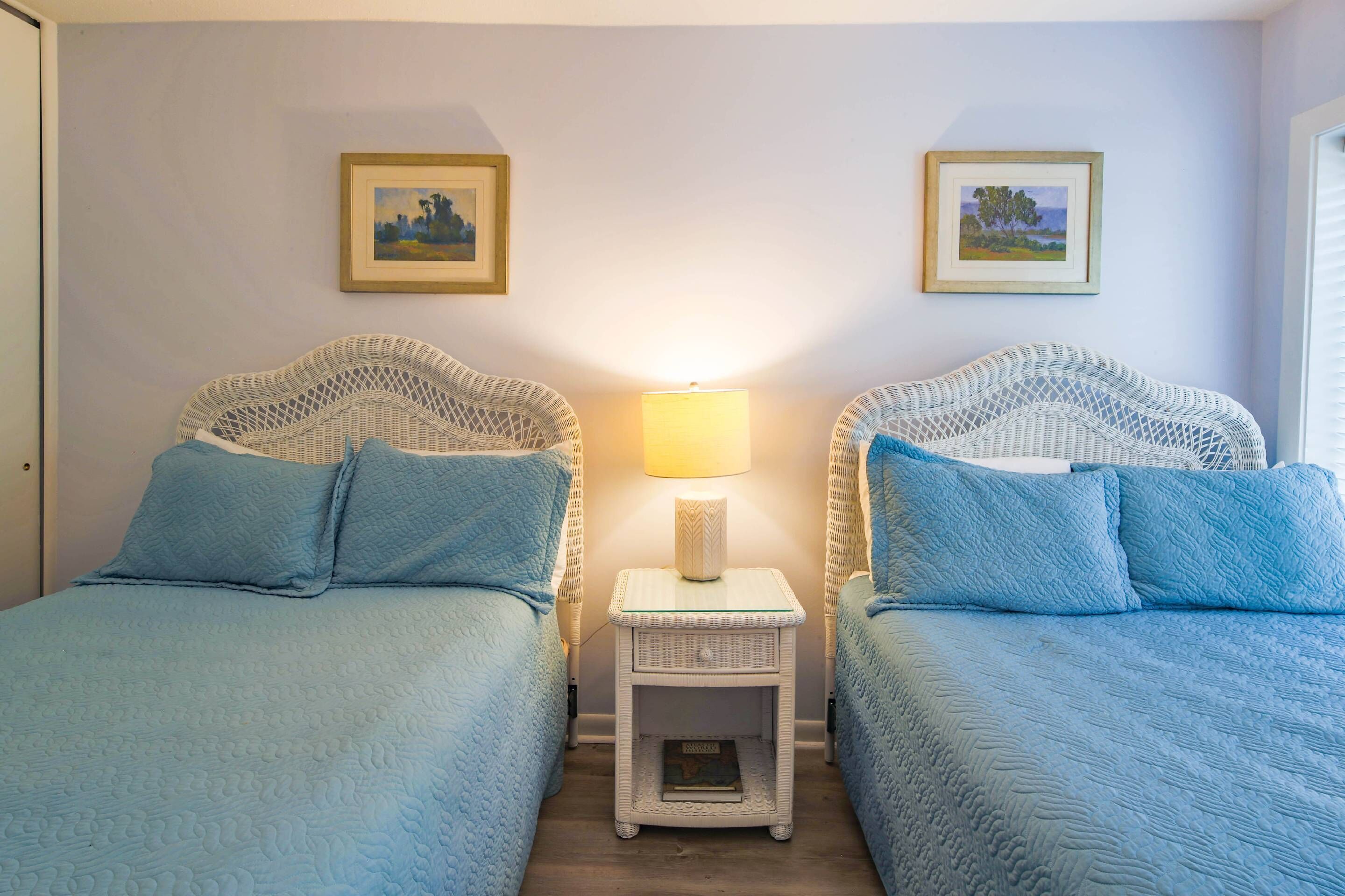 A room with two beds, each with a blue quilted bedspread and matching pillows. There is a small white nightstand with a lamp between the beds.