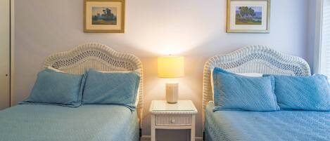 A room with two beds, each with a blue quilted bedspread and matching pillows. There is a small white nightstand with a lamp between the beds.