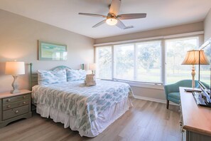 A bedroom with a bed, two bedside tables, a desk, a chair, a ceiling fan, and large windows.