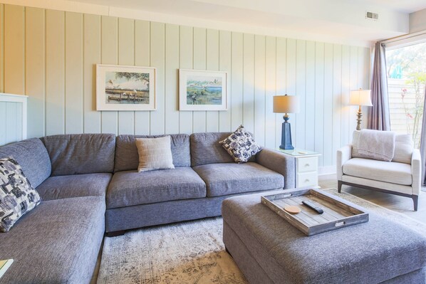 A living room with a grey sectional sofa, a small white side table, a lamp, and a chair. There are two framed pictures on the wall.