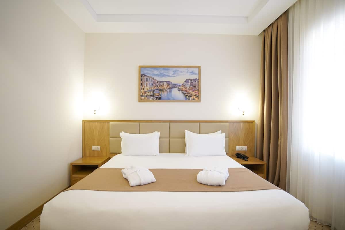 A hotel room with a large bed, two bedside tables, a headboard, a painting of a cityscape, and a window with curtains.