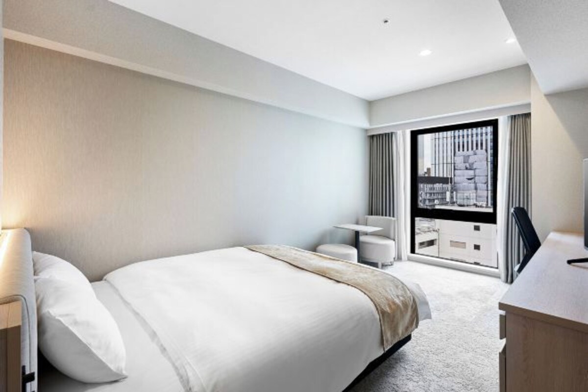 Una habitación de hotel moderna con una cama grande, un escritorio y vistas a la ciudad a través de ventanales de gran altura.