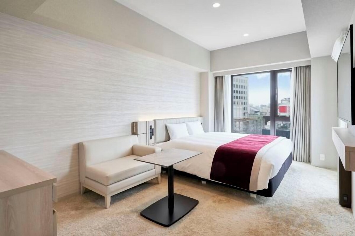 Una camera d'albergo con un letto, un divano, un tavolino, una lampada e vista sulla città attraverso ampie finestre.