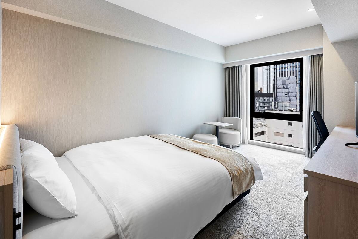 A modern hotel room with a large bed, a small desk, and a view of the city through a large window.
