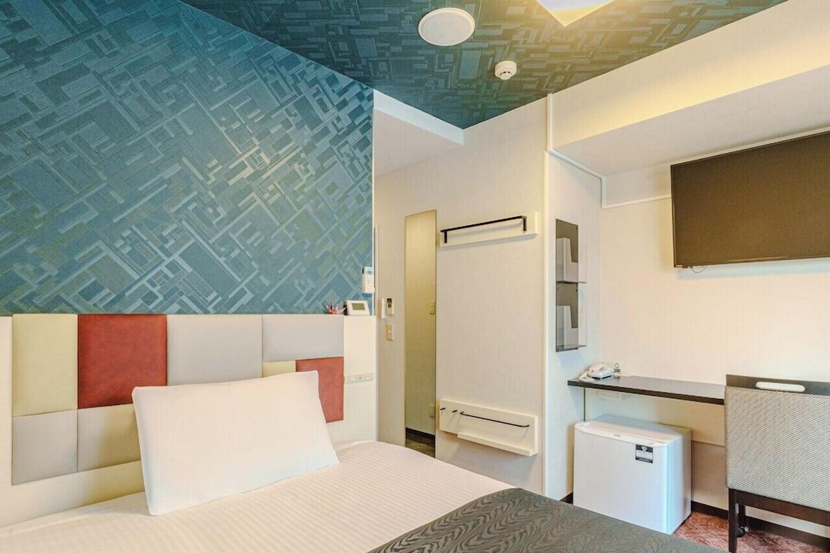 A hotel room with a bed, a desk, a chair, a small refrigerator, and a bathroom with a shower.
