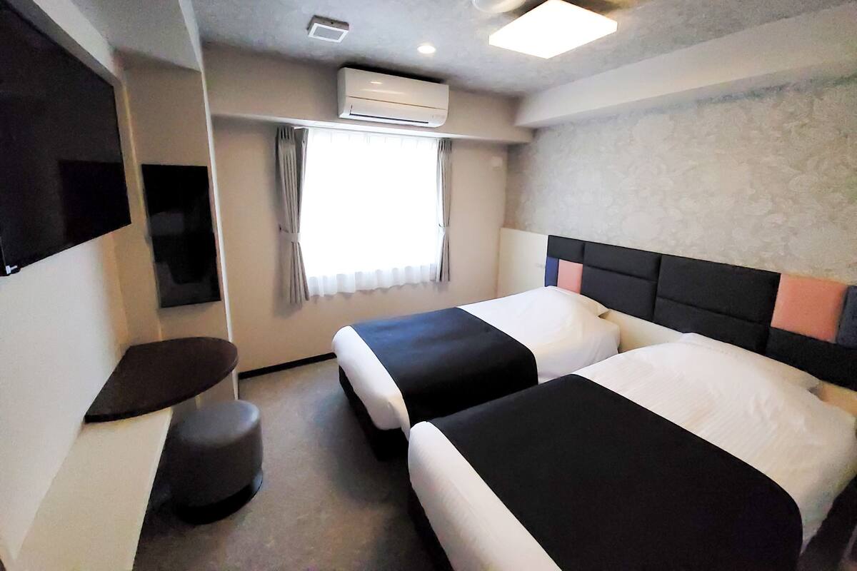 A hotel room with two beds, a TV, an air conditioning unit, and a window with curtains.