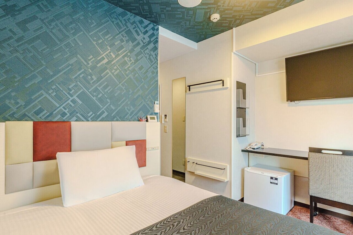 A hotel room with a bed, a desk, a small refrigerator, and a mirror.