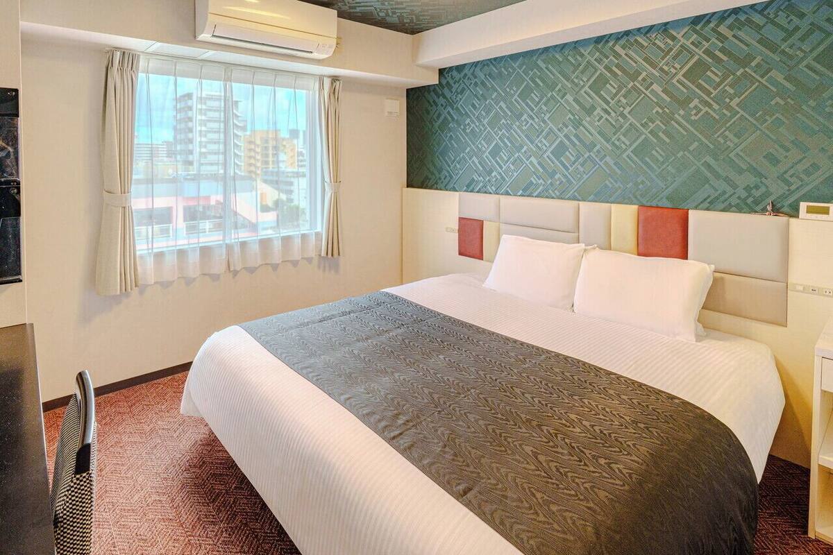 A hotel room with a large bed, a headboard with a geometric pattern, an air conditioning unit, a window with curtains, and a view of buildings outside.