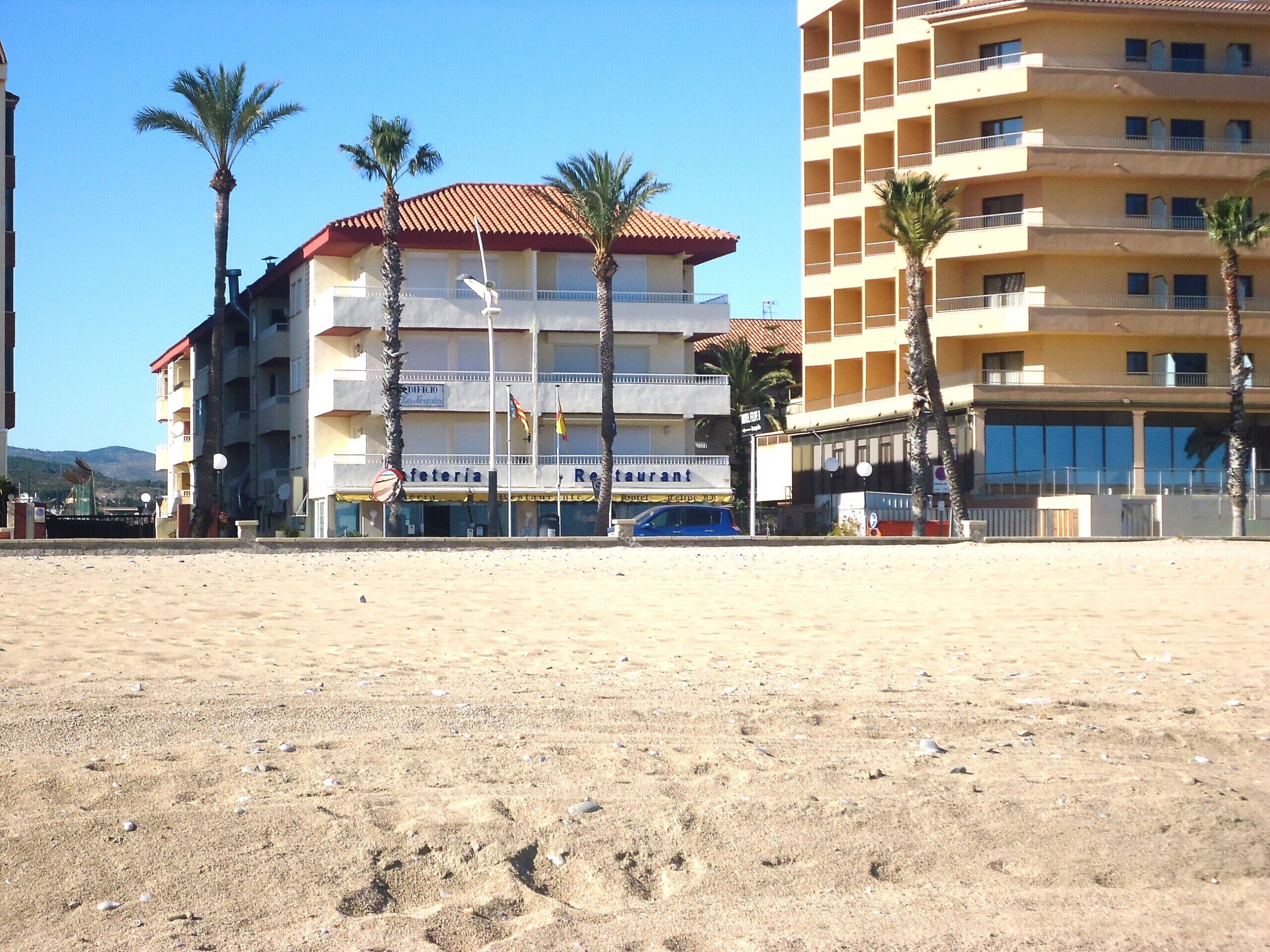 Ein mehrstöckiges Hotel mit Restaurant im Erdgeschoss, Palmen und einem Sandstrand im Vordergrund.