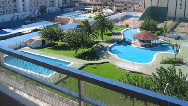 Un complejo hotelero con varias edificaciones, una piscina central y zonas verdes circundantes.