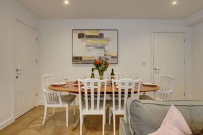A dining area with a wooden table, white chairs, and a painting on the wall. - Marston Croft Cottage | Hot Tub | Sleeps 7 |Southam | Inspire Homes (Southam)