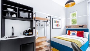 Kompakti hotellihuone, jossa on pieni keittiönurkkaus, värikkäällä tekstiilillä varustettu sänky, ikkuna, josta on näkymä, ja riippuvalaisin.