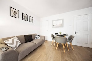 A modern living room with a grey sofa, a dining table with chairs, and framed botanical prints on the wall.