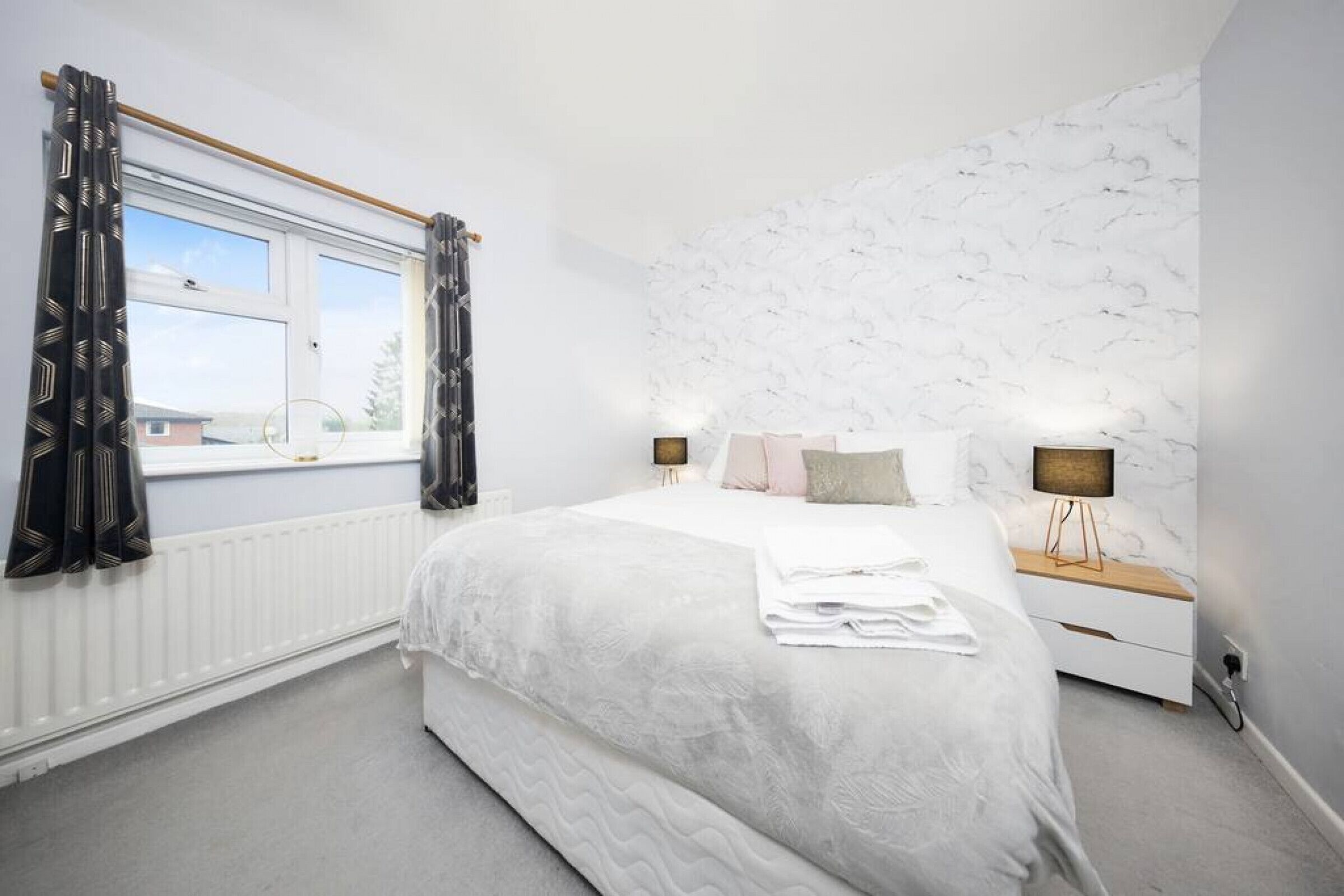 A bedroom with a large bed, a window with curtains, a nightstand with a lamp, and a marble-patterned wall.