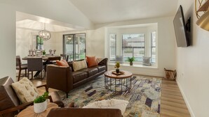 A spacious living room with a brown leather sofa, a coffee table, and a patterned rug. There is a dining area with a chandelier and a view of the outdoors through large windows. - Single-Level 3BR Scottsdale Home | Pool Table, Fire Pit (Scottsdale)