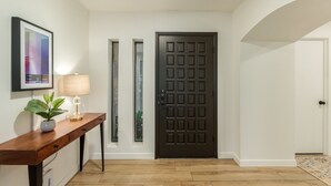 A hallway with a dark wooden door, a framed picture on the wall, a small table with a lamp and a plant, and a wooden floor. - Single-Level 3BR Scottsdale Home | Pool Table, Fire Pit (Scottsdale)