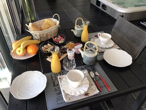 Ett frukostbord med ett varierat utbud av matvaror, inklusive frukt, bröd och drycker, uppdukat på ett svart bord med utsikt över en bubbelpool i bakgrunden.