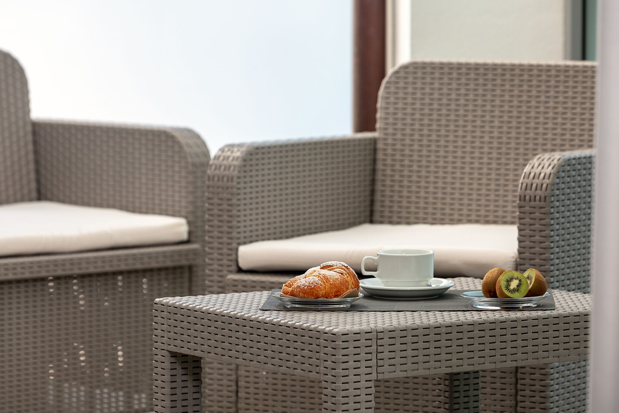 Móveis de vime com almofadas, um prato com um pastel e kiwi, e uma chávena sobre uma mesa.
